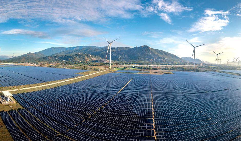 Energia Renovável — Aerial view of a vast solar farm with wind turbines against a mountainous backdrop on a sunny day.